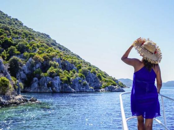 Frau im Blauen Kleid steht auf Boot und blickt auf Wasser und Küste der Türkei