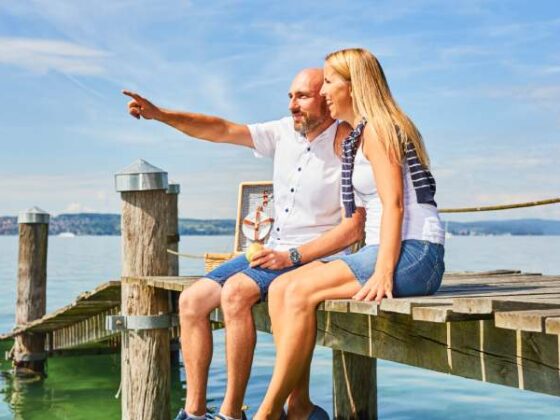 Paar sitzt am Steg am Bodensee und zeigt in die Ferne