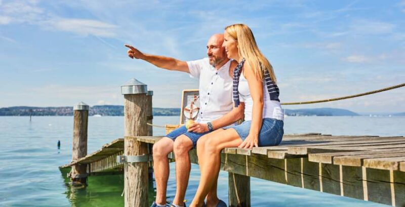 Paar sitzt am Steg am Bodensee und zeigt in die Ferne