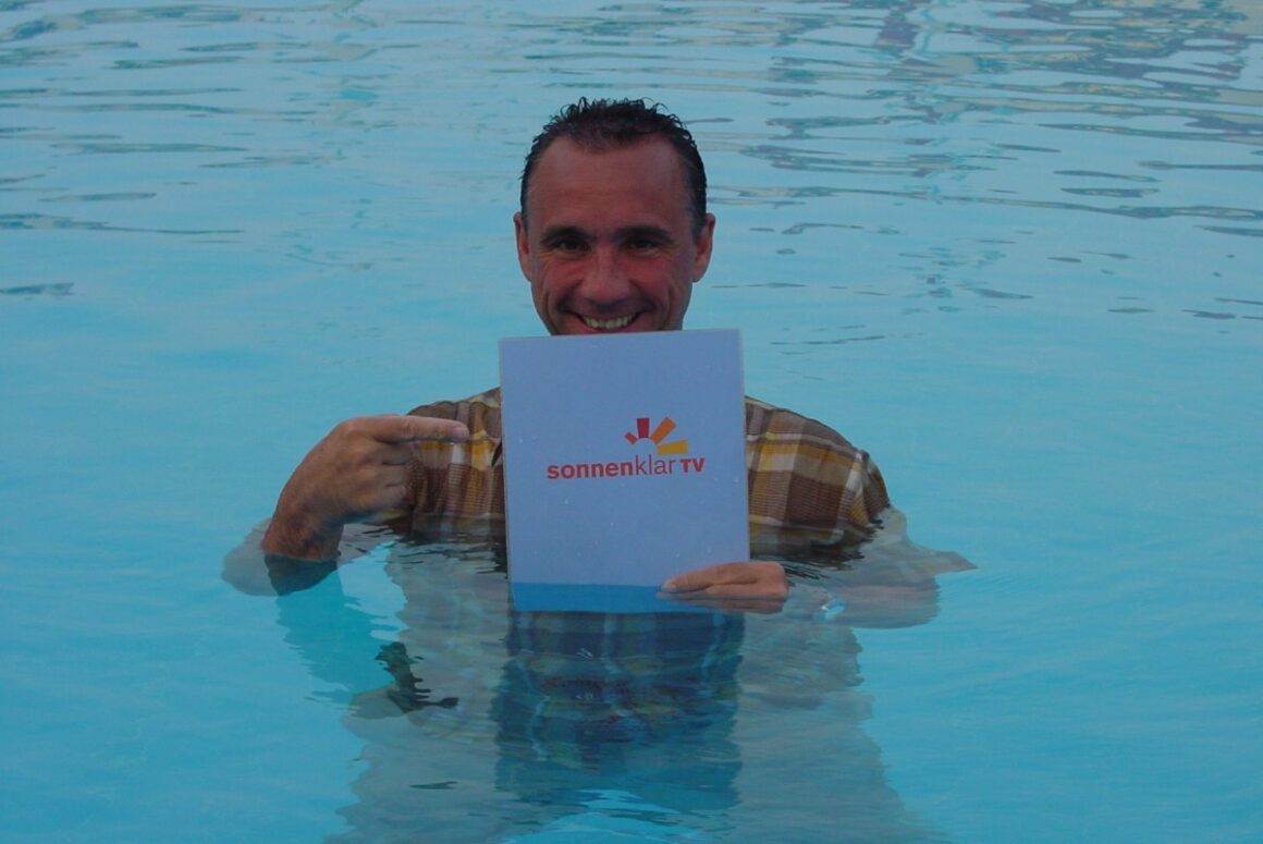 Ulf-Dieter Kunstmann in einem Pool, zeigt lachend auf seine sonnenklar.TV Moderationskarte