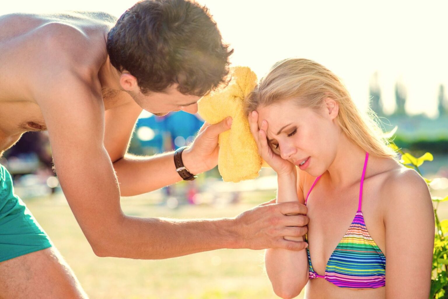 Krank im Urlaub – das sind eure Rechte und Pflichten im Überblick Frau am Strand mit Kopfschmerzen und Mann mit Handtuch hilft ihr