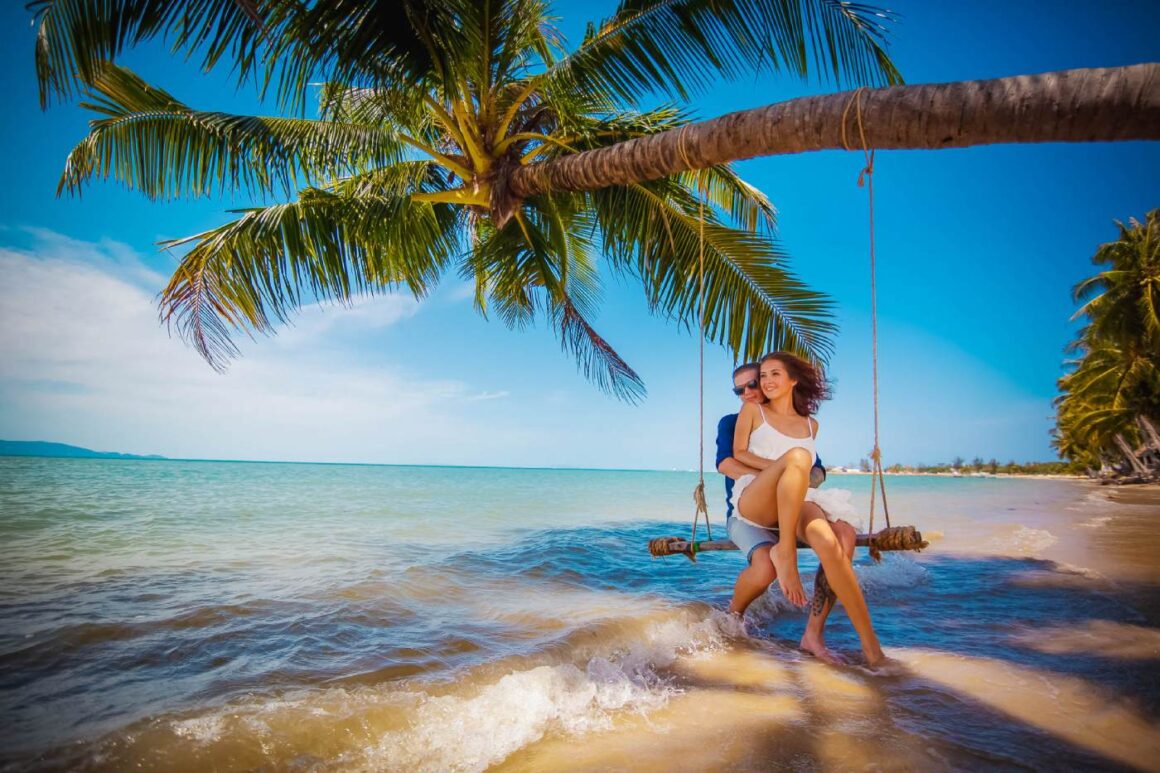 Günstigste Reisezeit 2027: Ferienfreie Zeiten in Deutschland Junges Paar an einem tropischen Strand auf einer Schaukel, die an einer Palme hängt