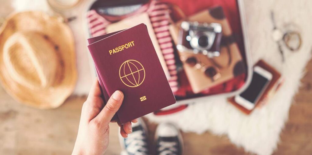 Hand hält Reisepass, im Hintergrund ein geöffneter Koffer und Strohhut