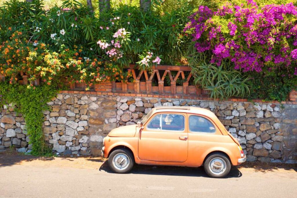 Ein Italienisches gelbes Auto in Taormina, Italien, parkt an einer mit Blumen bewachsenen Mauer
