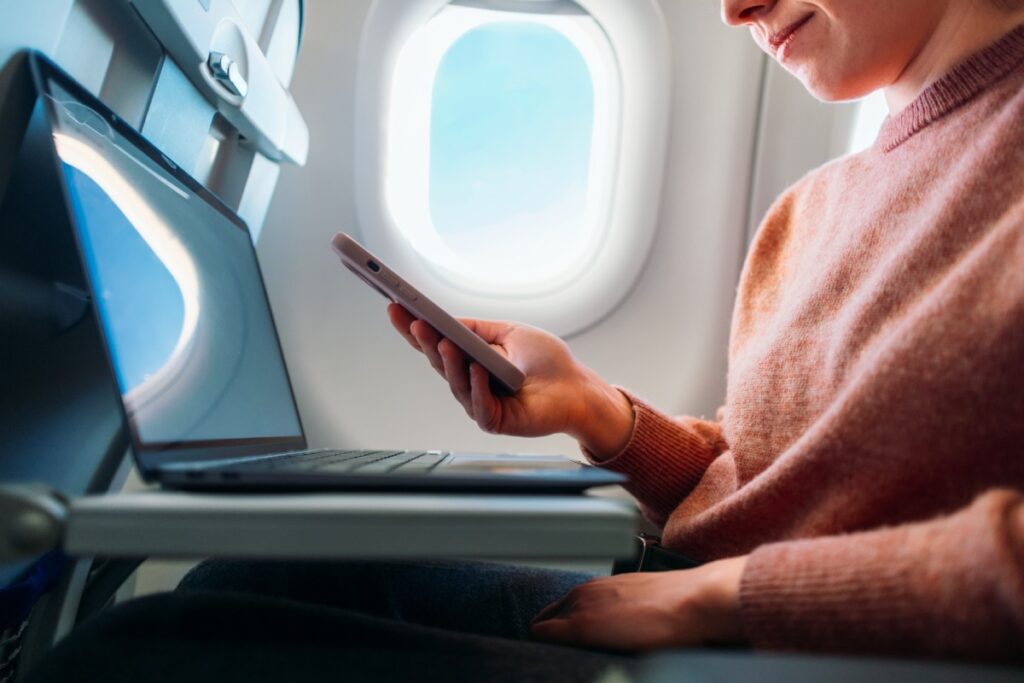 Person mit Smartphone in der Hand und Laptop im Flugzeug