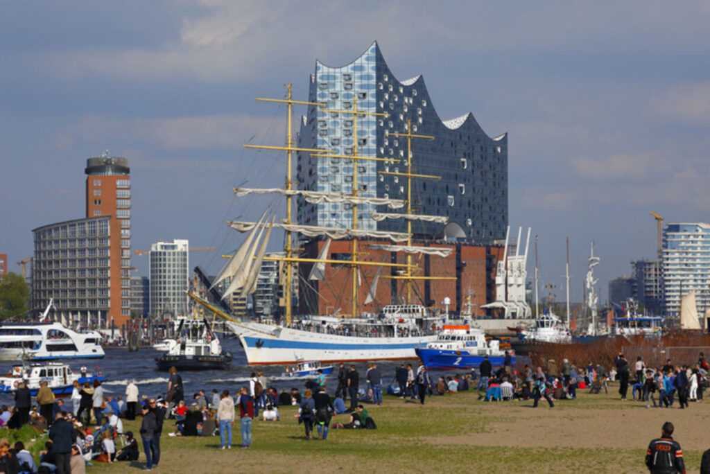 Menschen stehen am Ufer der Elbe mit Blick auf die Elbphilharmonie während ein Dreimaster vorbeifährt