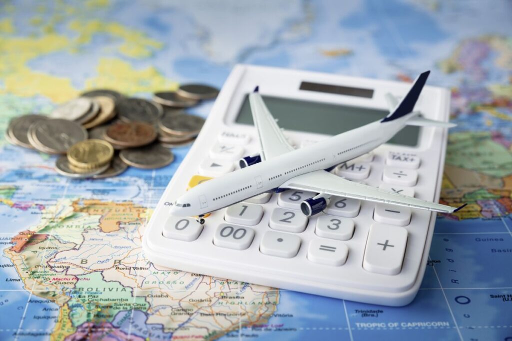 Modellflugzeug auf Taschenrechner mit Münzen auf Weltkarte als Symbol für steigende Reisekosten und Urlaubsbudget