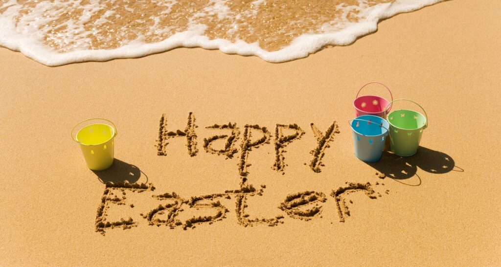 Von Stau bis Streik: Was Oster-Urlauber jetzt wissen müssen Happy Eastern in den Sand gemalt und bunte Eimer stehen am Strand