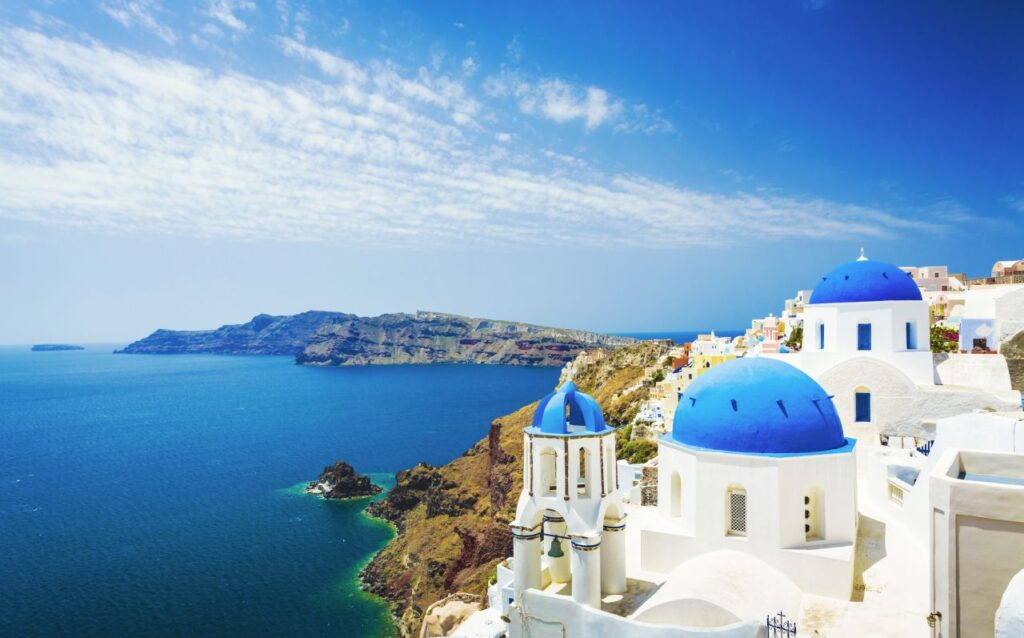 Griechenland-Preise steigen: Womit Reisende jetzt rechnen müssen Blick über die blau-weiß gemusterten Dächer der Kirche in Oia Stadt auf der Insel Santorin in Griechenland über das blaue Meer und die Felsenküste.