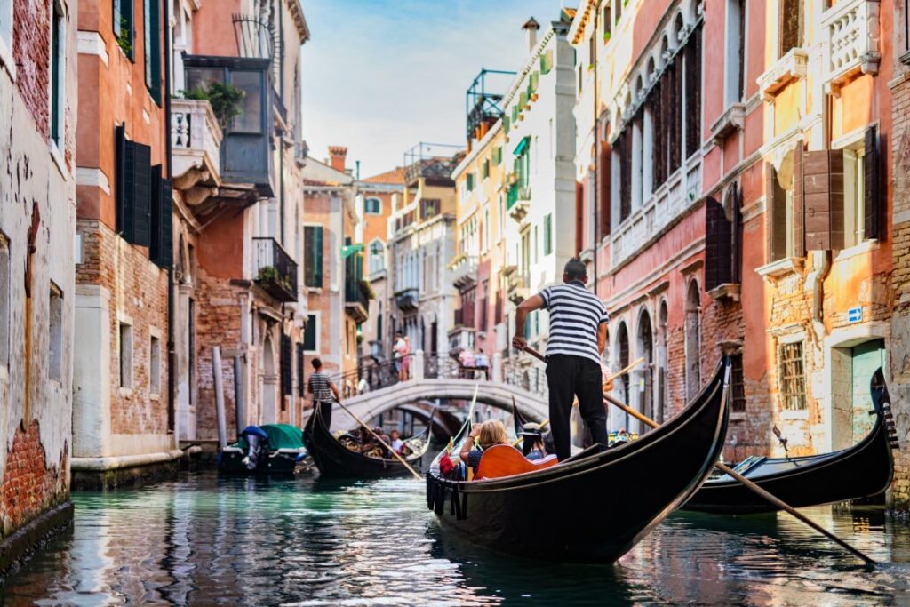 Gondeln mit Touristen und Gondoliere bei gutem Wetter unterwegs auf einem Kanal in Venedig mit Brücke und Häusern im Hintergrund