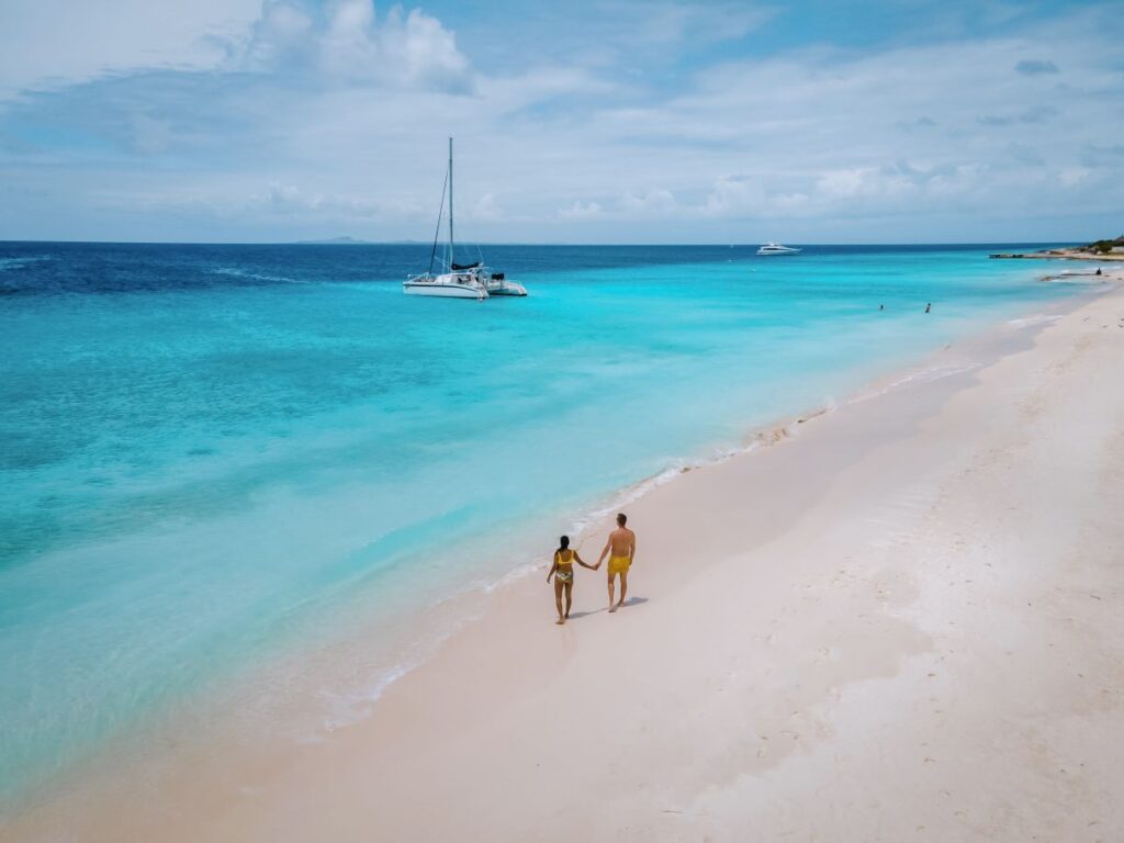 Direktflug nach Curaçao: Karibikinsel rückt für deutsche Urlauber näher Ein Paar läuft am puderweißen Sandstrand vor türkisblauer Meereskulisse und Katamaran auf der Karibikinsel Curaçao.
