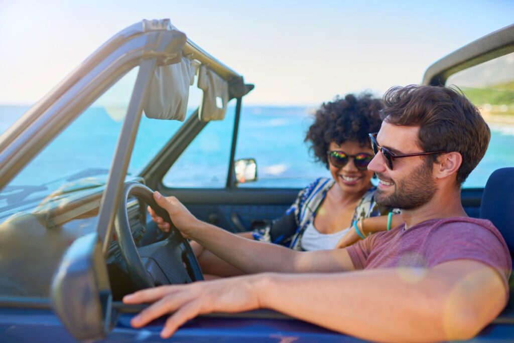 Stressfalle Mietwagen: So schützen sich Urlauber vor teuren Überraschungen Ein Paar - er am Steuer, sie ihn verträumt anschauend daneben - sitzt entspannt und lachend in einem Cabrio, das entlang der Küste vor blauer Meereskulisse fährt.