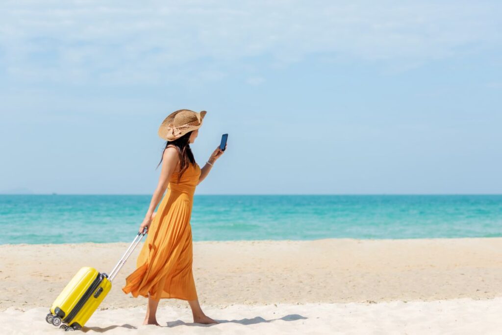 Handykosten im Urlaub: So vermeiden Reisende teure Roaming-Fallen Eine Frau im Sommerkleid und mit Hut, spaziert mit gelbem Rollkoffer an einem Sandstrand und checkt ihr Handy auf Nachrichten.