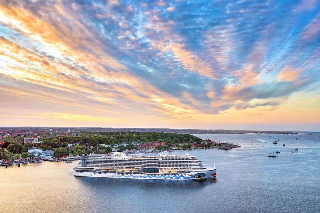 Ein Kreuzfahrtschiff von AIDA legt bei Sonnenuntergang und orange gefärbtem Wolkenhimmel von Kiel aus ab.