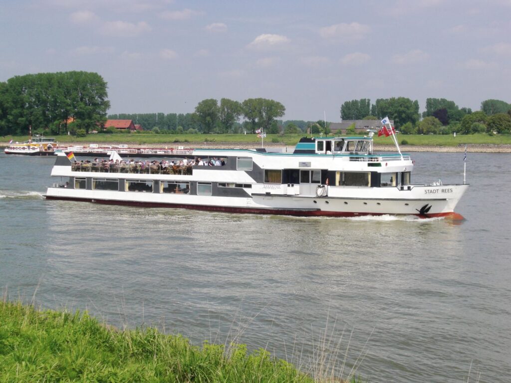 Flusskreuzfahrten unter Druck: Was das Niedrigwasser jetzt für Reisende bedeutet Ein Schiff der Stadt Rees mit Passgieren am Oberdeck auf dem Rhein entlang eines grünen Wiesenstücks.