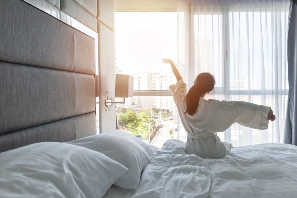 Eine Frau streckt sich am Morgen vorm Fenster in ihrem Hotelbett