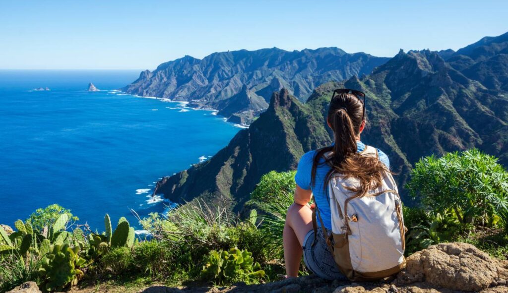 Singlereisen-Studie: Wer besonders gern allein in den Urlaub fährt Frau auf Wanderung blinkt von Berg aus auf Küste, Meer und blauen Himmel